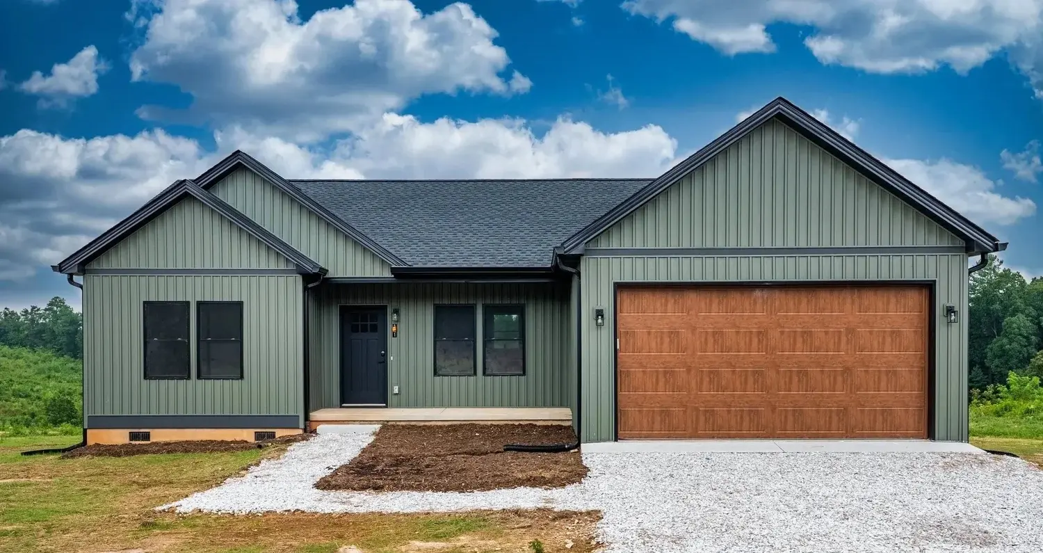 Front view of a new residential house with vertical green siding, a dark front door, and a two-car garage with a wooden door. The house is set against a partly cloudy sky with greenery surrounding it.