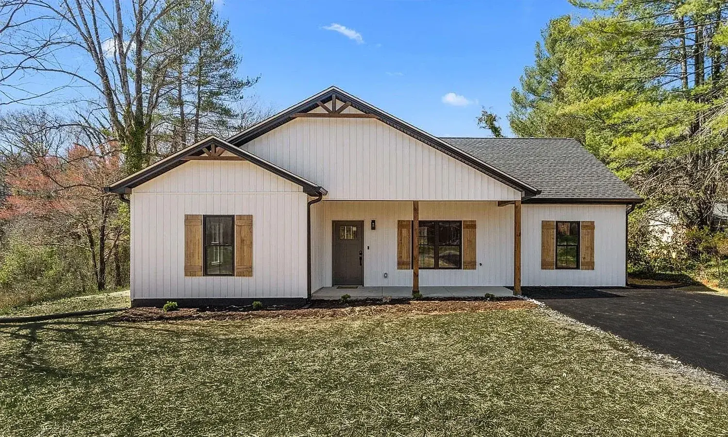 White single-story house with wooden shutters, front porch, and dark roof surrounded by trees.